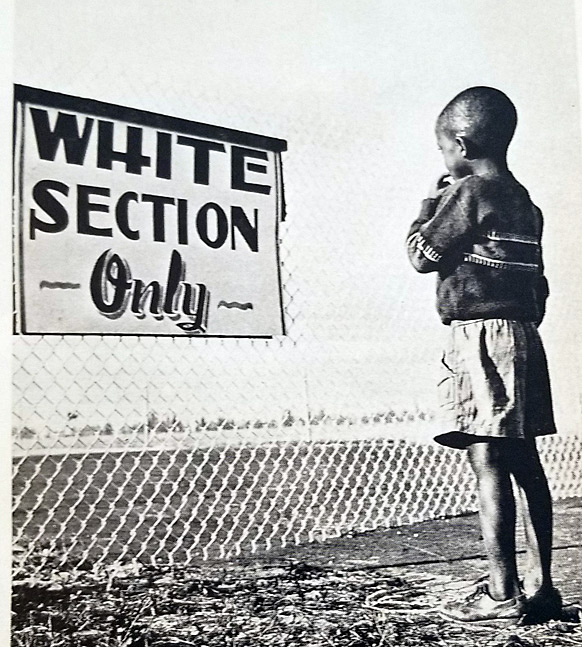 A black boy stands in front of a sign that bars him from what looks like a ball field.